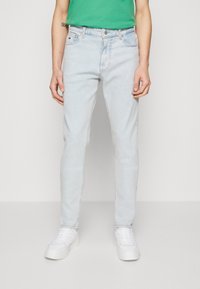 Jeans in denim blu chiaro con una vestibilità slim, dotati di zip sul davanti, passanti per cintura e tasche anteriori. Abbinati a sneakers bianche e una camicia verde.