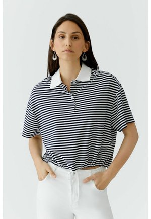 Frau mit langen dunklen Haaren, die ein schwarz-weiß gestreiftes kurzärmeliges Poloshirt mit weißem Kragen und weiße Hose trägt, Hände in den Taschen.