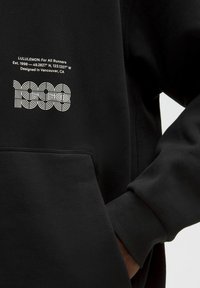 Czarna bluza z kapturem wykonana z miękkiego materiału, z kieszenią z przodu. Białe nadrukowane napisy zawierają "LULULEMON" i "1998" w abstrakcyjnym wzorze.