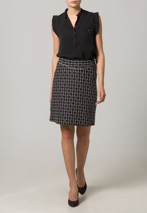 Kobieta w czarnej, bezrękawnikowej bluzce, czarnej spódnicy do kolan z białym geometrycznym wzorem i czarnych szpilkach na szarym tle.
