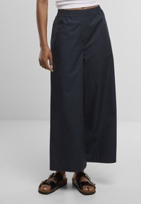 Personne portant un pantalon noir large et ample et des sandales noires à bout ouvert avec de grosses boucles, debout sur un fond uni.