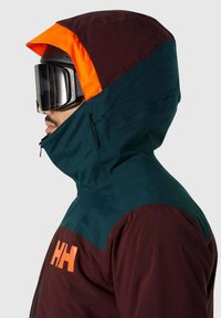 Skijacke mit einem kastanienbraunen Körper, grünen Schultern und einer orangefarbenen Kapuze. Glatte Textur, mit einer seitlichen Reißverschlusstasche und einer großen Skibrille.