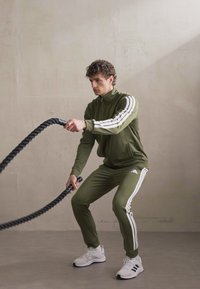Olíva zöld szövetből készült sportos tréningruha, fehér csíkokkal az ujjakon és az oldalán, fehér sportcipőkkel párosítva, vastag fekete kötelet tartva.