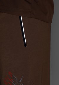 Pantalon marron avec une poche latérale bordée de noir et blanc, avec de petits détails graphiques rouges et gris près du bas.