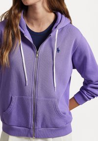 Felpa viola con zip, dotata di cordini bianchi, tasche frontali e un piccolo logo. Il tessuto ha una consistenza morbida ed è caratterizzata da una vestibilità casual.