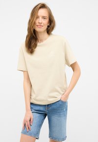 Beige cotton t-shirt with a round neckline and short sleeves, featuring a small white shell graphic on the chest, paired with light denim shorts.