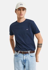 T-shirt in cotone blu navy con scollo rotondo, maniche corte e piccolo logo ricamato sul petto; abbinata a jeans azzurri e una cintura marrone.