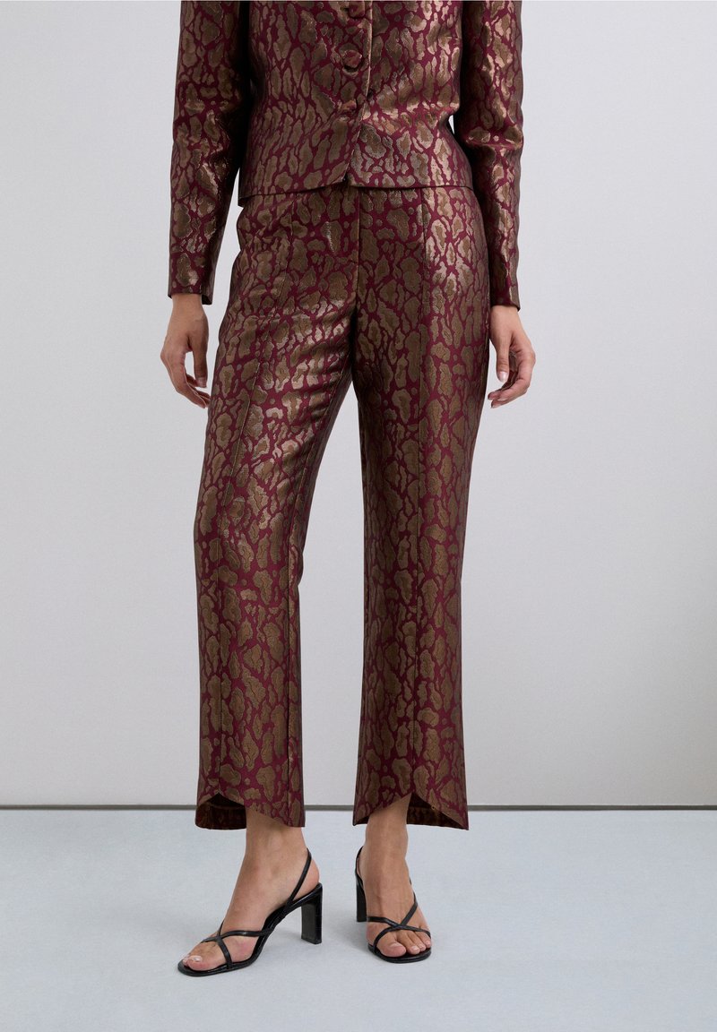 Mujer vistiendo pantalones metálicos burdeos con un patrón dorado y chaqueta a juego, con sandalias de tacón alto negras con tirantes, sobre un suelo gris claro.