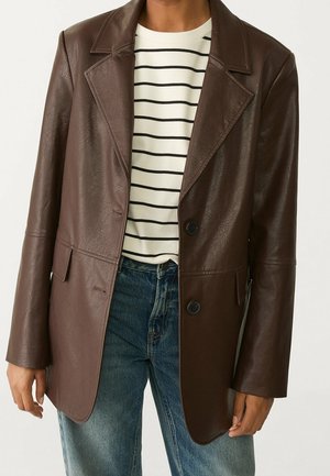 Persoon draagt een donkerbruine leren blazer over een wit overhemd met zwarte horizontale strepen en een blauwe spijkerbroek.