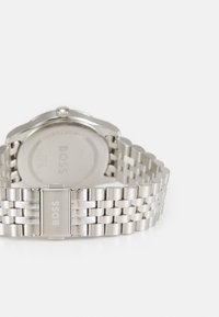 Montre en acier inoxydable argenté avec un bracelet à maillons, présentant un boîtier rond minimaliste et une finition mate lisse. Logo affiché sur le fermoir.