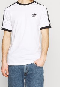 Bílý tričko s černými akcenty, kulatým výstřihem a logem Adidas na levé straně hrudi. Krátké rukávy s černými pruhy.