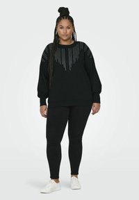 Černá mikina se strukturovaným designem a flitry ve tvaru V na přední straně, spárovaná s černými legínami a bílými teniskami.
