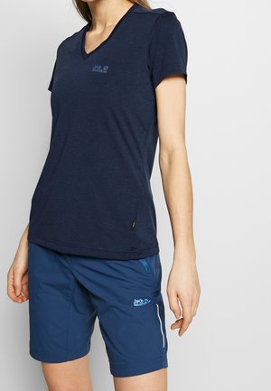 Osoba nosząca granatowy t-shirt Jack Wolfskin z dekoltem w serek i pasujące niebieskie krótkie spodenki outdoor z zamykaną kieszenią i logo na udzie.