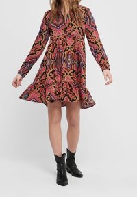 Robe à manches longues avec un motif paisley audacieux présentant du rose, du bleu et de l'or sur un fond noir, avec un ourlet évasé. Des bottines noires aux chevilles sont portées.