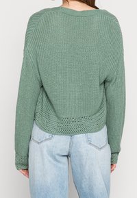Žena v volné pletené svetru v šalvějově zelené barvě s otevřenými detaily na manžetách a lemu, spárovaného se světle modrými džíny s vysokým pasem.