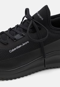 Černé tenisky s vrchní částí z meshového materiálu a gumovou podešví. Mají ploché tkaničky, detaily s logem a strukturovaný design pro přilnavost a flexibilitu.