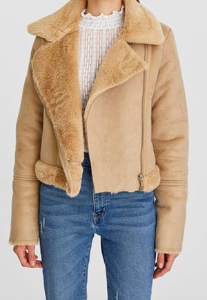 Veste en shearling beige avec grand col, fermeture éclair asymétrique et doublure en fausse fourrure portée sur un chemisier blanc texturé et un jean bleu.