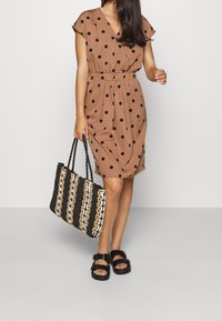 Ruskea pilkkudressi, jossa on lyhyet hihat, vyötä ympäröivä vyötärö ja polveen ulottuva helma. Kudottu musta, valkoinen ja ruskea laukku. Mustat sandaalit solkiin.