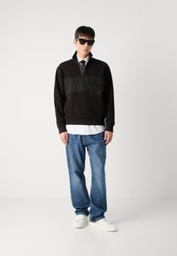 Felpa nera con design a mezza zip e tasca frontale, abbinata a una camicia bianca, jeans blu e sneaker bianchi. Tessuto testurizzato, fit casual.
