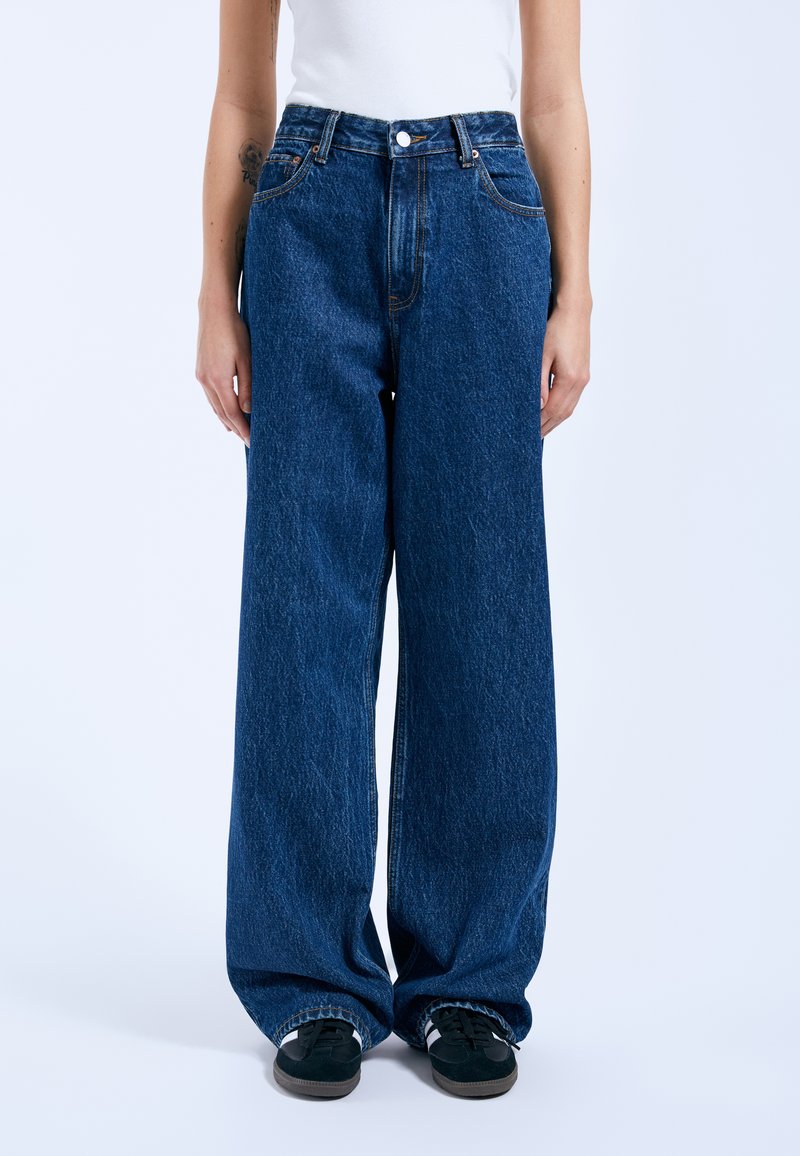 Weite Jeans aus blauem Denim mit hoher Taille, Vordertaschen und sichtbaren Nahtdetails. Kombiniert mit schwarzen Sneakers.