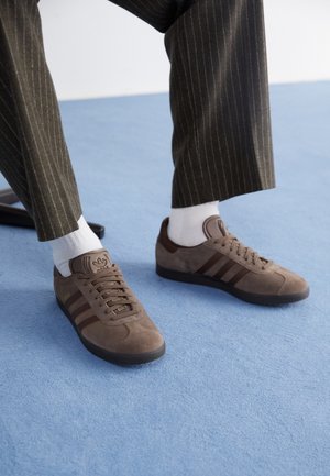 Osoba ubrana w brązowe zamszowe trampki Adidas Gazelle, białe skarpetki i spodnie w prążki, stojąca na niebieskim dywanie.