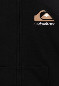 Sweat à zip noir fabriqué en tissu doux. Présente un logo Quiksilver marron et blanc sur le coin supérieur droit. Comprend des poches avant et une finition texturée.
