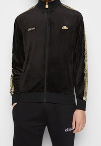 Svart velour träningsoverall med dragkedja i framkant, med guld accenter och logotyp på bröstet, ribbade ärmslut och matchande svarta byxor.