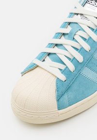 Blå mockasinsneakers med en texturerad krämfärgad gummitå och vita snören. Utmärkande sömnadsdetaljer längs sidorna och snörningsområdet.