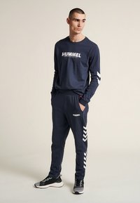 Marinblå långärmad topp med "HUMMEL"-logotyp, kombinerad med matchande joggers med chevron-accenter och luftig passform. Textilen verkar slät.
