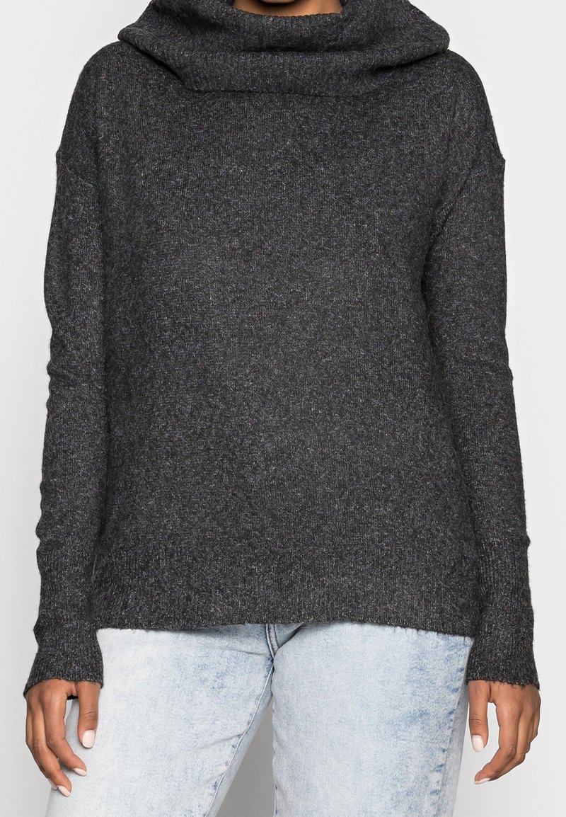 Kolgrå tröja med cowl-hals, ribbad textur och långa ärmar. Har en avslappnad passform och något bred nederkant, kombinerad med ljusblå jeans.