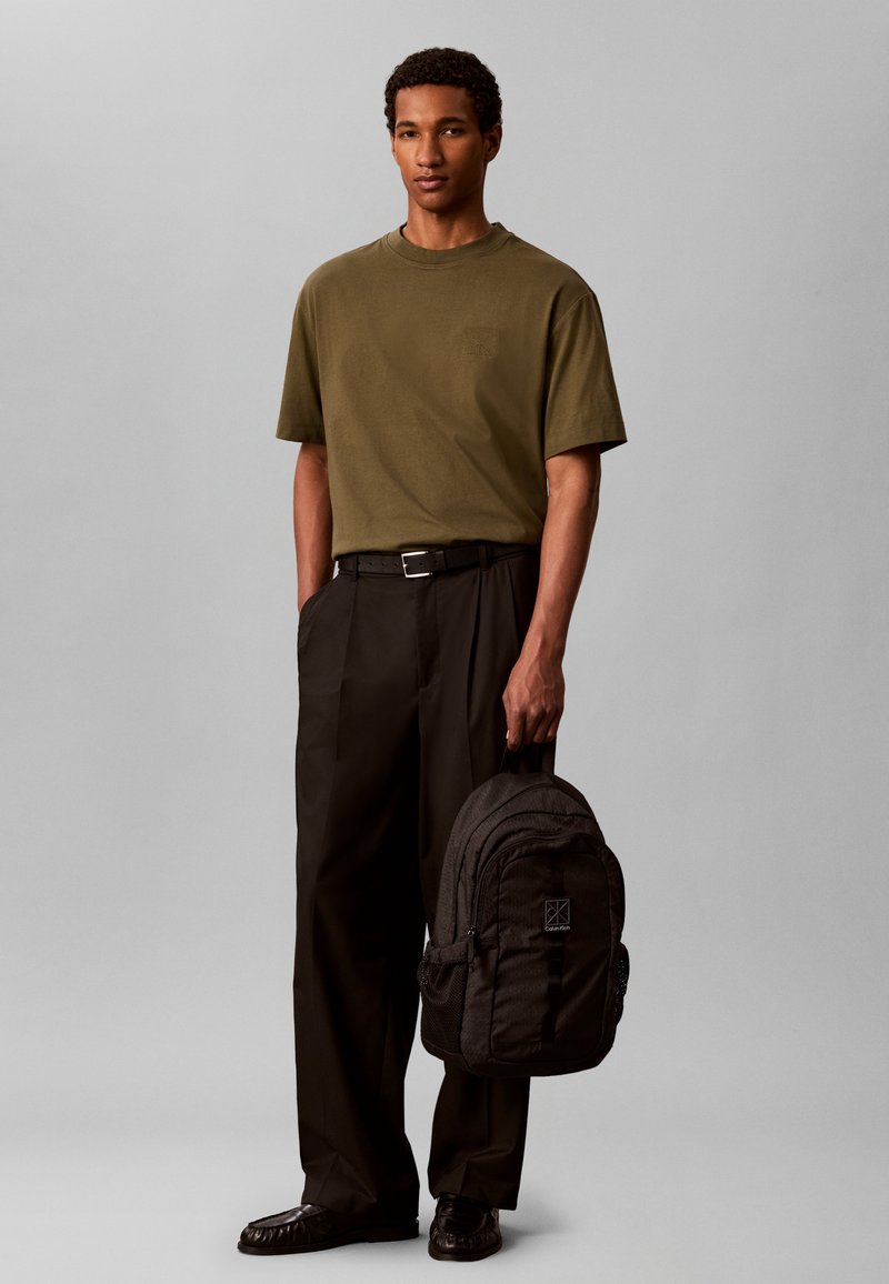 Jeune homme debout portant un t-shirt vert olive, un pantalon noir, des chaussures noires et tenant un sac à dos noir dans sa main droite.