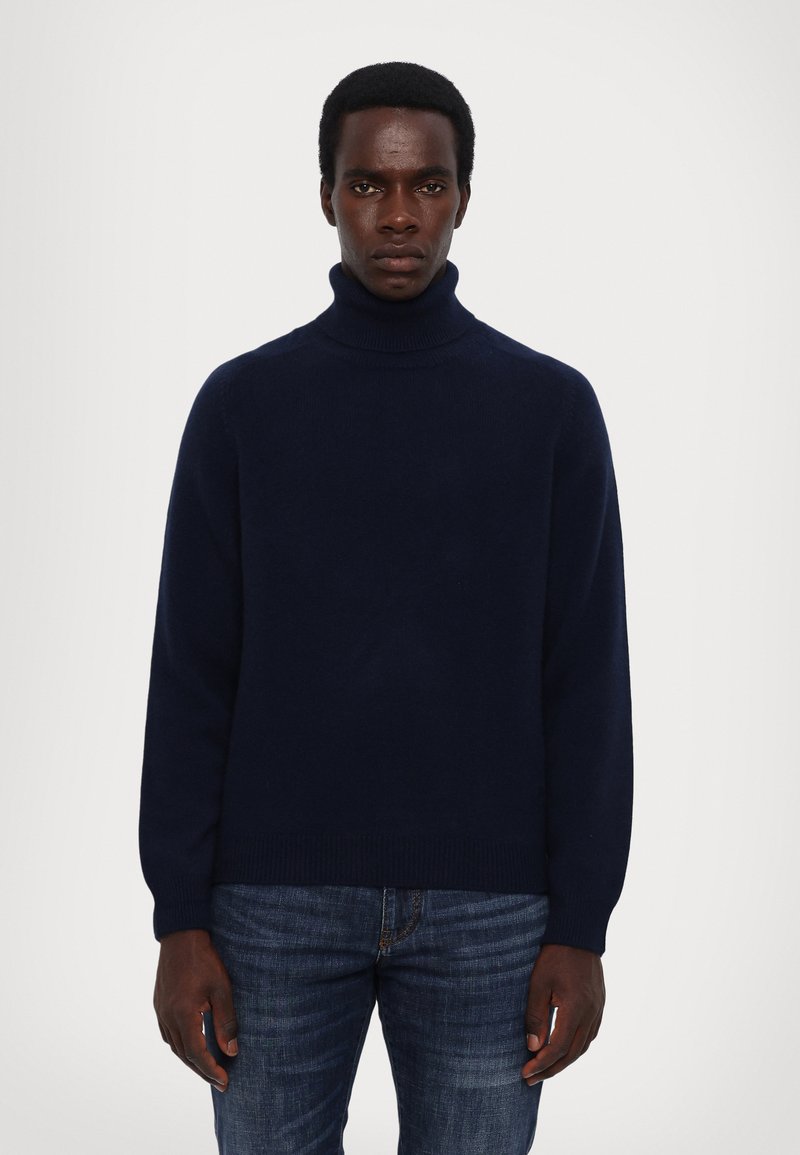 Navy turtleneck sweater in soft knit fabric, featuring long sleeves, ribbed cuffs, and a relaxed fit, worn with blue denim jeans.