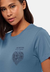 Blaues Baumwoll-T-Shirt mit einem Herzdesign, das mit aufwendigen Mustern gefüllt ist, einschließlich Schmetterlingen, und dem Text: "JACK WOLFSKIN WIR LEBEN, UM ZU ENTDECKEN."