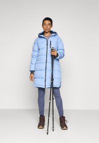 Piumino azzurro chiaro con cappuccio, due tasche laterali, chiusura con zip, indossato su leggins grigi e stivaletti marroni, persona che tiene bastoni da trekking.