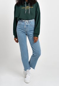 Sudadera verde con logo dorado, combinada con jeans ligeros de tiro alto de corte recto y dobladillo deshilachado, llevada con zapatillas blancas.