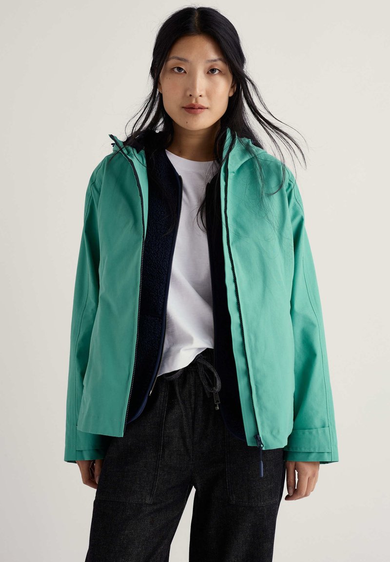 Veste imperméable bleu sarcelle avec col montant, fermeture éclair et doublure intérieure en peluche, associée à un t-shirt blanc et un pantalon foncé.
