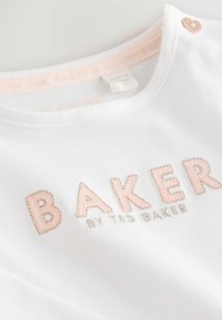 Valkoinen puuvillainen t-paita, jossa on vaaleanpunaisella ja metallinhohtoisella hopealla brodeerattu "BAKER". Pyöreä kaula-aukko, jossa on vaaleanpunainen yksityiskohta ja sydämen muotoinen nappi.