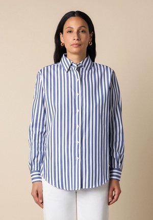 Vrouw die een blauw-wit verticaal gestreept overhemd met knopen en een witte broek draagt, staand tegen een effen beige achtergrond.