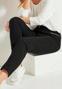 Schwarze, figurbetonte Hose mit Seiteneingriffen und glatter Textur, kombiniert mit einem weißen Langarmoberteil. Die Schuhe sind weiße Sneaker mit beigen Akzenten.