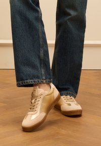 Des baskets beiges avec des accents dorés métalliques, des surfaces en daim et lisses, une semelle en gomme texturée, associées à un jean en denim sombre, coupé au-dessus de la cheville.