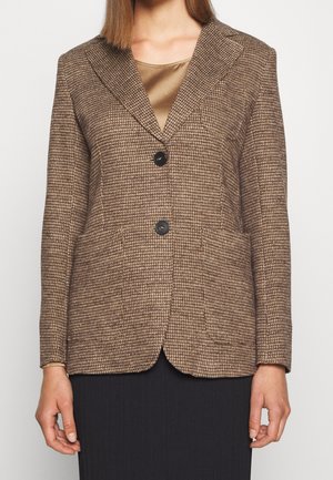 Vrouw draagt een bruin geruit colbert met twee zwarte knopen over een satijnen zandkleurige top en een zwarte rok.