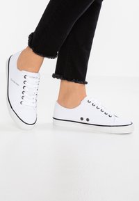 Weiße Leinwand-Sneaker mit schwarzen Akzenten, ausgestattet mit einer schwarzen Gummisohle, runder Spitze und einem Schnürdesign. Logo an der Seite aufgedruckt.