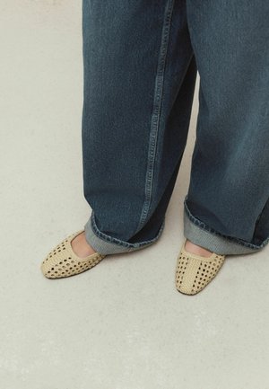 Legs in loose dark blue jeans cuffed at the bottom paired with beige woven slip-on shoes on a light floor.