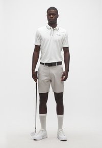 Homme portant un polo blanc, un short beige et des baskets blanches, tenant un club de golf, debout devant un fond blanc uni.