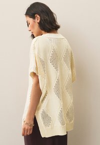 Kremasto obarvan pulover na pletenje s kratkimi rokavi, diamantnimi vzorci in odprtimi pletenimi deli. Ima rob z rebrastim zaključkom in ovratnik.