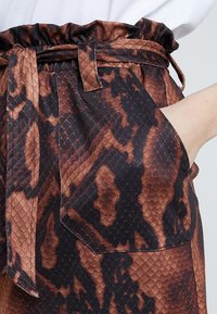 Kalhoty s potiskem hnědých a černých hadích šupin, se staženým pasem, páskem, kapsami a texturovanou tkaninou napodobujícími šupiny plazů.
