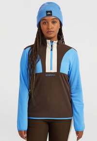Bruine en blauwe fleece pullover met rits en hoge kraag, voorpaneel in het wit en O'Neill-logo; model draagt ook een blauwe beanie.