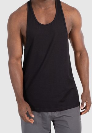 Muskelöser Mensch trägt ein schwarzes ärmelloses Tanktop und graue Shorts, steht mit entspannten Armen an den Seiten.