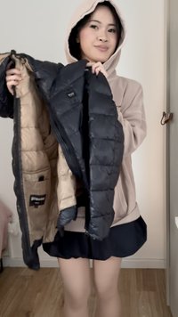 Žena oblečená v béžovej mikine s kapucňou a čiernej sukni drží otvorenú čiernu prešívanú bundu s béžovým vnútrom vnútri miestnosti.