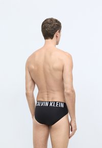 Crne kupaće gaće imaju široki pojas s natpisom "CALVIN KLEIN" u bijeloj boji, od glatke tkanine, i uski su fit oko tijela.